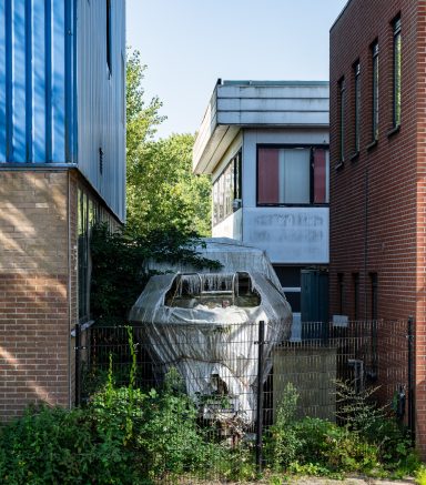 Een verwaarloosde boot tussen gebouwen, omringd door groen en een hek.